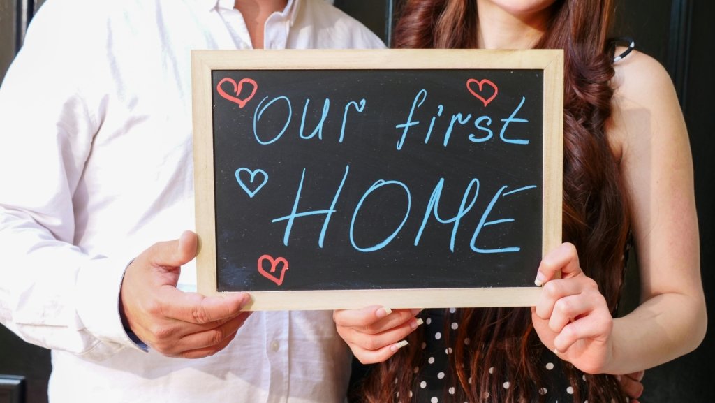 Couple holding a chalkboard sign that says "Our First Home" with hand-drawn hearts.