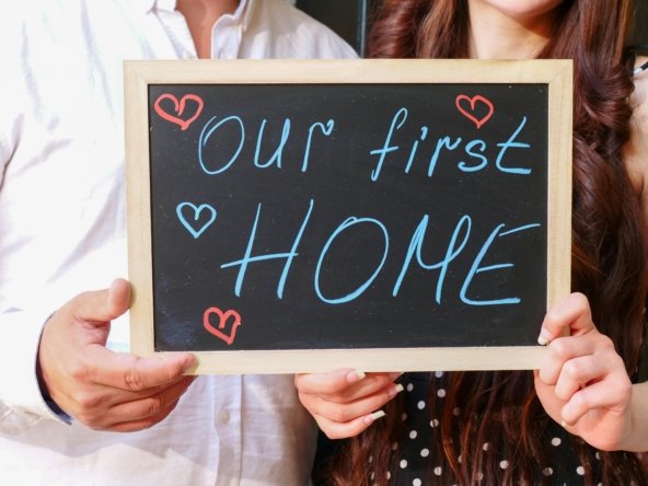 Couple holding a chalkboard sign that says "Our First Home" with hand-drawn hearts.