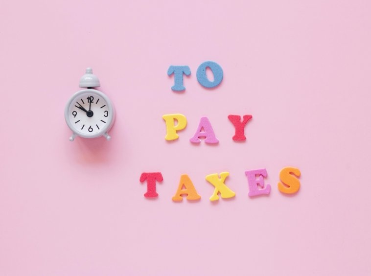Colorful foam letters spelling “TO PAY TAXES” next to a white alarm clock, all set against a pink background.